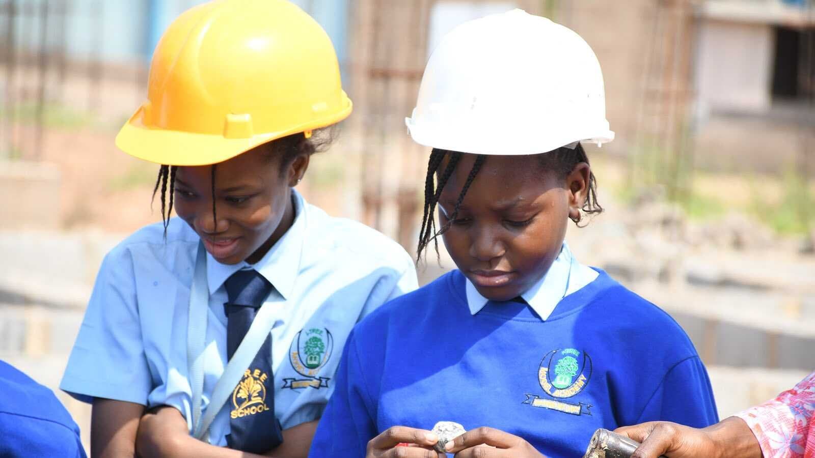 Two Nigerian students learning practical skills together, wearing blue school uniforms and safety helmets